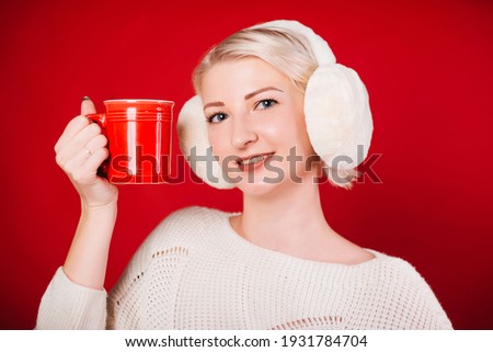 A woman in a white sweater with headphones on a red background holds a mug of coffee in her hands. Studio photo