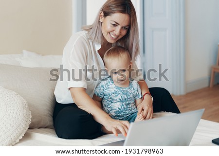 Young mother with her child working on laptop in bedroom at home. Multi-tasking, freelance and motherhood concept