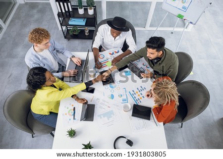 Competent IT specialists in stylish outfit working on modern gadgets while sitting together at bright office. Young multiracial people shaking hands, talking and smiling during cooperation. Royalty-Free Stock Photo #1931803805
