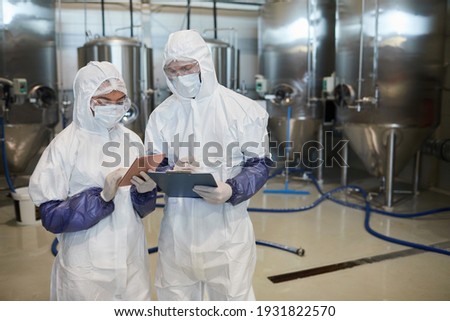 Waist up portrait of two young workers wearing hazmat suits while using digital tablet at chemical factory, copy space Royalty-Free Stock Photo #1931822570