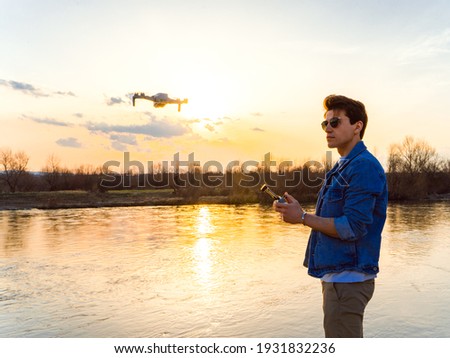 handsome young man with the sunglasses stands by the river flying a drone holding the controller in his hands, trying to capture an amazing colour of the sky
