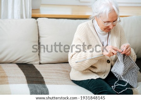 Closeup of senior woman, engaged in knitting sitting on the sofa at home. She has gray hair and hyperopia glasses Royalty-Free Stock Photo #1931848145