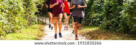 A group of high school cross country runners running on a dirt path in a park surrounded by bushes and trees. Royalty-Free Stock Photo #1931850266