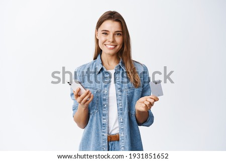 Portrait of good-looking smiling woman with blond hair, holding plastic credit card with smartphone, paying at online shopping application, purchase product, white background