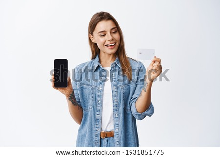 Online shopping. Smiling millennial female model showing empty smartphone screen with plastic credit card, mobile banking app conept, white background
