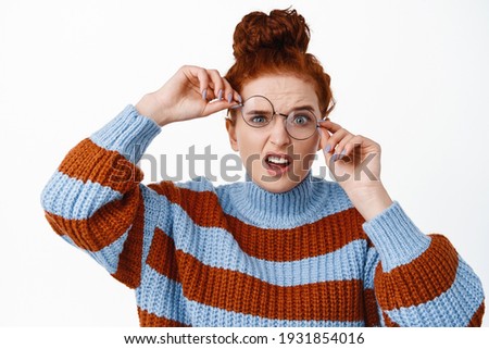 Young nerdy girl with ginger hair and blue eyes, trying to wear crooked broken glasses, cant see without eyewear, standing funny against white background