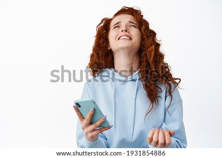 God why me. Miserable and sad redhead girl complain on unfair and cruel life, holding smartphone, raising head up and whining as losing online, standing against white background Royalty-Free Stock Photo #1931854886