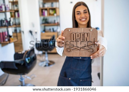 Young woman opening a beauty store and looking very happy - small business concepts. Smiling owner of hair salon standing with sign open and leaning on glass door