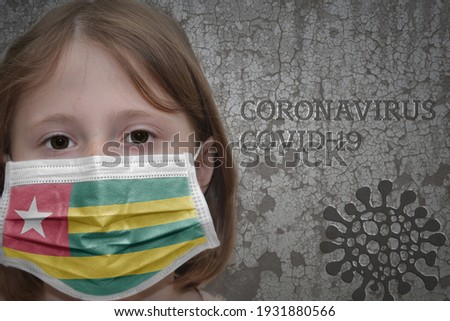 Little girl in medical mask with flag of togo stands near the old vintage wall with text coronavirus, covid, and virus picture. Stop virus