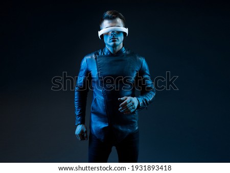 Young man in futuristic costume on dark background. Guy using VR helmet. Augmented reality, virtual reality, future technology, game concept. Blue neon light. 