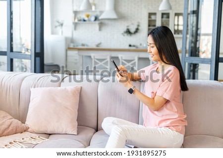 Pretty Asian lady with long black hair, sitting on the couch, looking at cellphone, video-calling, texting, talking to friend, family, chatting on social media, refreshing memory looking at old photos