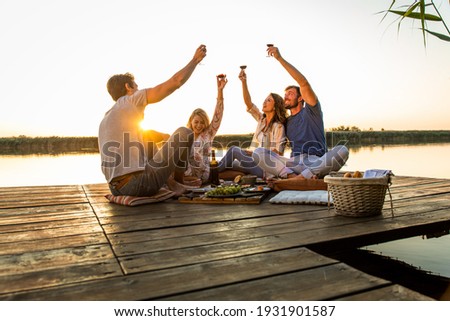 Group of friends having fun on picnic near a lake, sitting on pier eating and drinking wine. Royalty-Free Stock Photo #1931901587