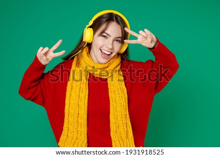 Cheerful young brunette woman 20s wearing basic casual knitted red sweater yellow scarf listening music with headphones showing victory sign isolated on bright green color background studio portrait
