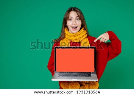 Excited young brunette woman 20s in basic knitted red sweater yellow scarf point index finger on laptop pc computer with blank empty screen isolated on bright green color background studio portrait