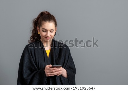curly-haired emotional young woman in a black cloak using mobile phone, isolated on gray background