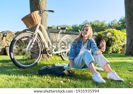 Rest, leisure, lifestyle. Young woman relaxing sitting on grass in park using smartphone, bike background