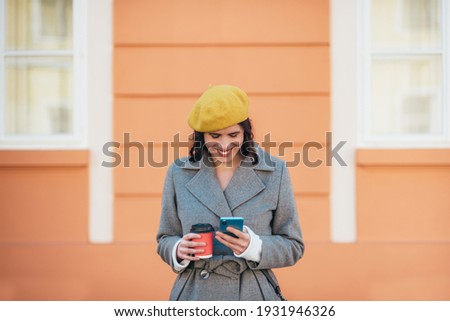 Portrait of an attractive young woman in gray coat wearing a yellow beret and using a smartphone while standing against the orange wall