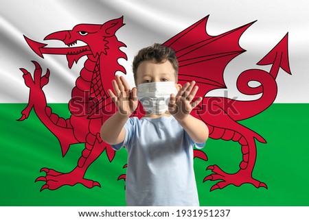 Little white boy in a protective mask on the background of the flag of Wales. Makes a stop sign with his hands, stay at home Wales.