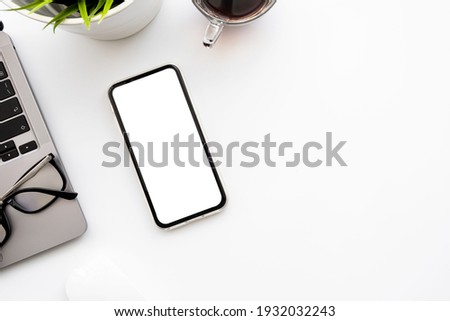 Smartphone with blank mockup screen is on top of white office desk table with laptop computer and office supplies. Top view with copy space, flat lay.