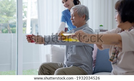 Young older senior asia citizen couple sit on sofa couch with in-home nursing care, assisted living, scrubs nurse use resistance band exercise for senior patient in physiotherapy treatment at home. Royalty-Free Stock Photo #1932034526