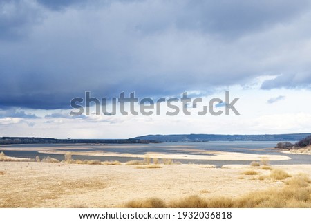 Picturesque golden autumn valley with bare trees on bank of large tranquil lake under sky with heavy grey clouds on nasty day