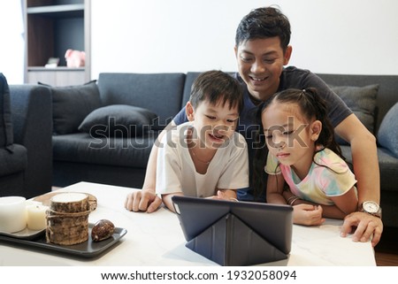 Positive smiling father and his two kids enjoying spending time together and watching educational show on digital tablet