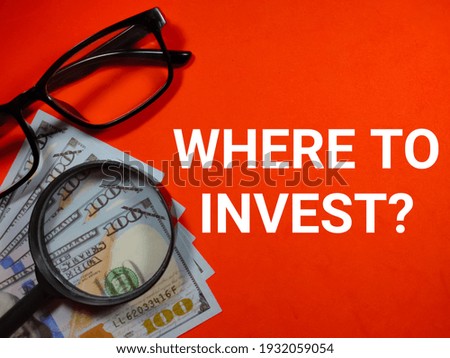Dollar banknotes,magnifying glass and glasses with text WHERE TO INVEST on red background.Business concept.
