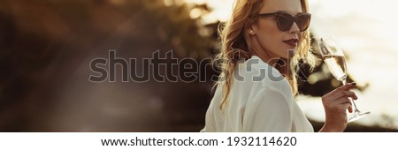 Stylish woman in sunglasses with a glass of wine outdoors. Elegant female drinking wine looking at camera. Large copy space in background. Royalty-Free Stock Photo #1932114620