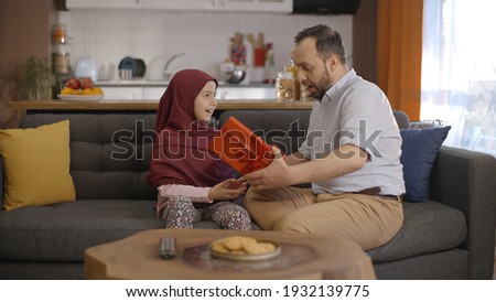 The affectionate little girl in hijab gives the gift box she received to her father on father's day. Young father hugs and kisses his little daughter. 