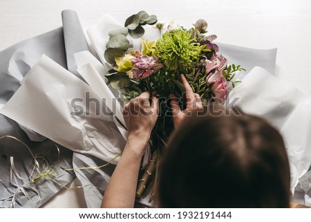 The hands of woman florist making a bouquet of mixed unusual original flowers in gray paper on a white background. Chrysanthemum green, yellow orchid flower, pink rose and green leaves.