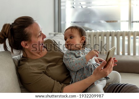 Happy mother and cute baby daughter enjoying using smartphone on sofa, smiling mum having fun with little girl playing mobile games watch funny videos. Baby girl with mom using phone together at home.