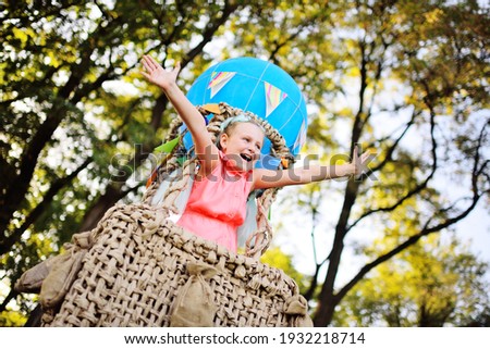 a little girl in pink clothes smiles sitting in a basket of a blue balloon against a background of sunlight and greenery.