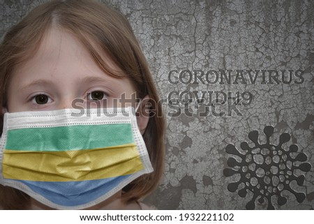 Little girl in medical mask with flag of gabon stands near the old vintage wall with text coronavirus, covid, and virus picture. Stop virus