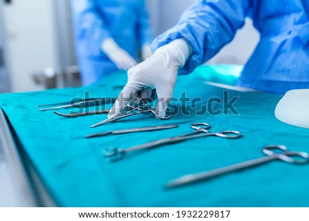 Nurse hand taking surgical instrument for group of surgeons at background operating patient in surgical theatre. Steel medical instruments ready to be used. Surgery and emergency concept Royalty-Free Stock Photo #1932229817