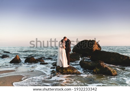 the groom and the bride stand on a stone in the sea at sunset. beautiful wedding couple at sunset of the day is on the beach. beautiful beach with gentle waves during sunset with newlyweds in the back Royalty-Free Stock Photo #1932233342
