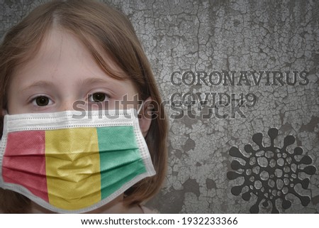 Little girl in medical mask with flag of guinea stands near the old vintage wall with text coronavirus, covid, and virus picture. Stop virus