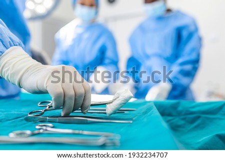 Nurse hand taking surgical instrument for group of surgeons at background operating patient in surgical theatre. Steel medical instruments ready to be used. Surgery and emergency concept Royalty-Free Stock Photo #1932234707