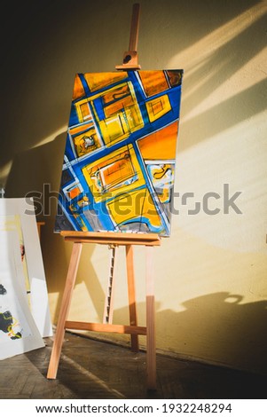 Easel with abstract geometric blue and orange painting in artist's workshop.