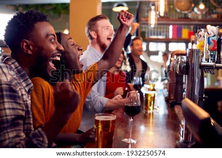 Group Of Excited Customers In Sports Bar Watching Sporting Event On Television Royalty-Free Stock Photo #1932250574