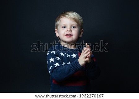 Boy in sweater with USA american flag claps his hands on black background with copy space.