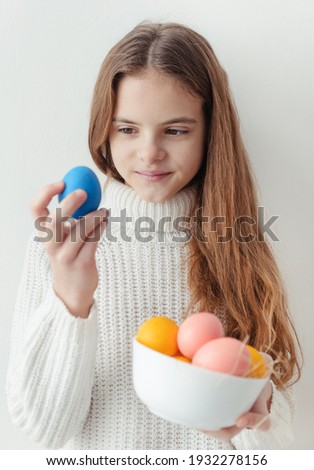 1 white cute European girl 10 years old with blue yellow and pink Easter eggs in a white sweater on a white wall background