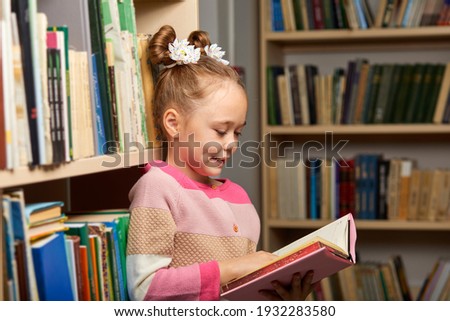 child girl get carried away by reading a book in library, standing betweel multi coloured bookshelves