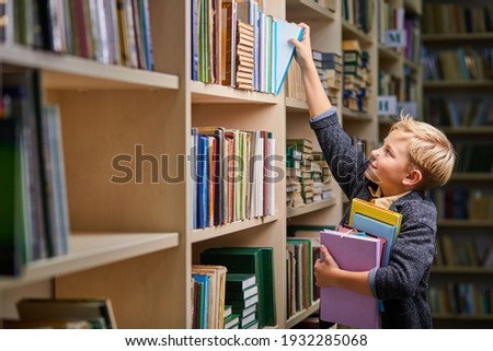 school boy taking books from shelves in library, with a stack of books in hands. child brain development, learn to read, cognitive skills concept