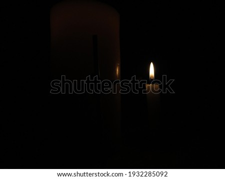 An Image of a white wax candle with a bright flame and a cross in the background in the dark.