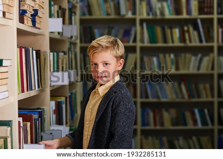 kid boy in library, looking at camera. school boy stand between shelves with books