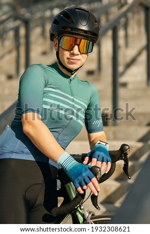 Portrait of young professional woman cyclist in cycling garment and protective gear posing for camera, standing on the steps with her bike while training outdoors