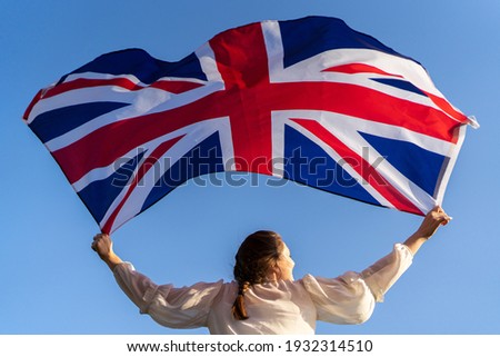 The flag of England waving in the hands of a woman against the blue sky. View from the back. Great Britain, victory and success Royalty-Free Stock Photo #1932314510