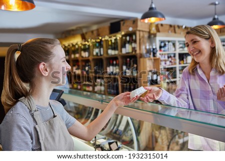 Female Customer Delicatessen Food Store Buying Local Cheese From Teenage Sales Assistant Royalty-Free Stock Photo #1932315014