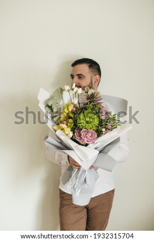 Beautiful young man in a white shirt holding a bouquet of mixed unusual original flowers in gray paper on a white background. Chrysanthemum green, yellow orchid flower, pink rose and green leaves.