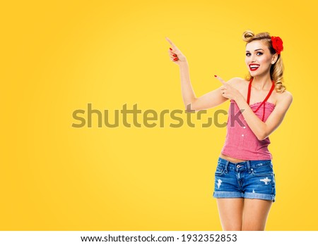 Amazed happy woman pointing at something. Excited girl in pin up, showing product or copy space for text. Retro fashion and vintage. Isolated over yellow color background. 